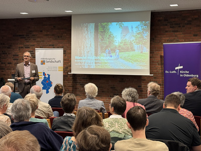 Im Vordergrund Publikum von hinten zu sehen, dieses schaut auf Pfarrer Stefan Welz, der im Hintergrund neben einer Leinwand steht.