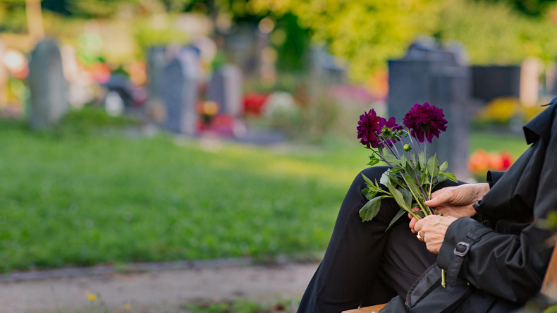 Ein Besucher eines Friedhofs sitzt und hält einige Blumen in der Hand. Im unscharfen Hintergrund sind Grabsteine zu erkennen.