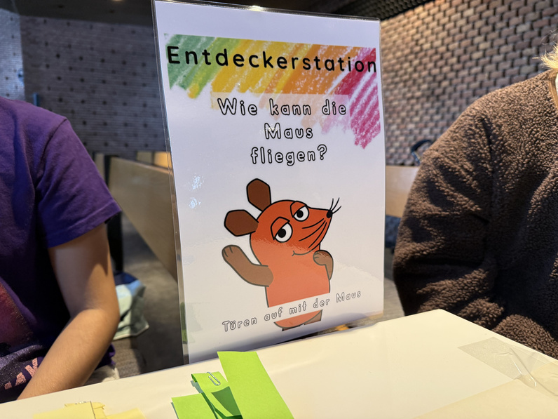 Auf einem bunten Schild steht die Frage "Wie kann die Maus fliegen?"