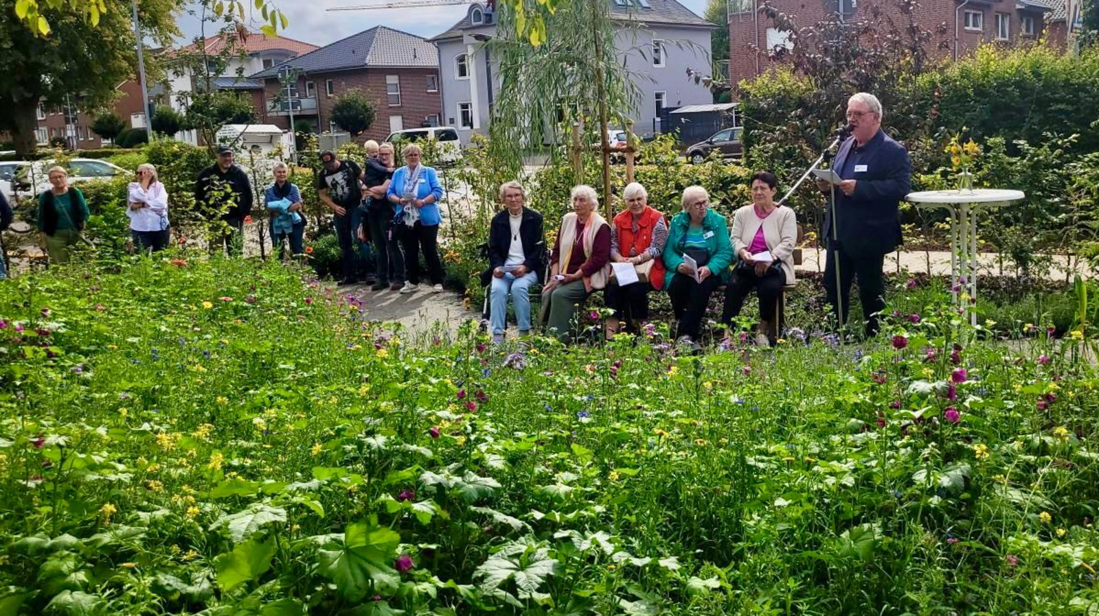 Menschen sitzen im Grünen auf Bänken, rechts im Bild steht Hans-Jürgen Hoffmann am Mikrofon und spricht.