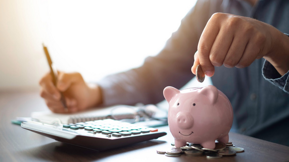 Ein Mensch sitz am Schreibtisch. Auf dem Schreibtisch liegen Unterlagen, ein Taschenrechner und ein Sparschwein. Mit der rechten Hand schreibt die Person in den Unterlagen und mit der rechten Hand wirft sie eine Münze in das Sparschwein.