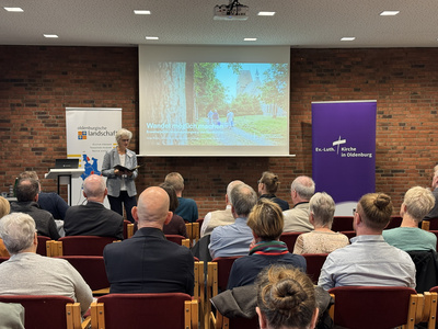 Im Vordergrund sitzt das Publikum, es ist von hinten zu sehen und schaut auf Oberkirchenrätin Gudrun Mawick, die neben einer Leinwand steht und spricht.