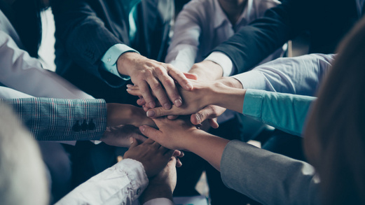 Ein Gruppe an Menschen steht im Kreis. Die Menschen strecken die Arme in die Mitte des Kreises und legen ihre Hände übereinander.