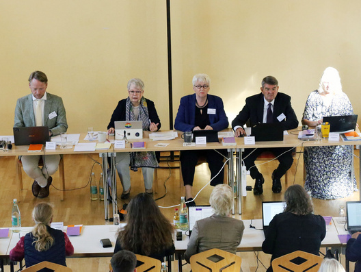 Die fünf Menschen des Präsidiums sitzen in einer Reihe an Tischen.