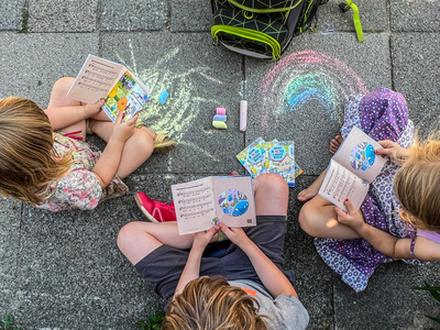 Drei Kinder sitzen auf dem Bürgersteig zwischen Kreide und Schulranzen und schauen in der "AchtKinderliederbuch"