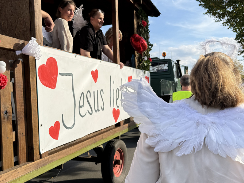 Link im Bild ist der Festwagen der Kirchengemeinde zu sehen, rechts im Bild eine Ehrenamtliche mit Engelsflügeln und Heiligenschein von hinten.