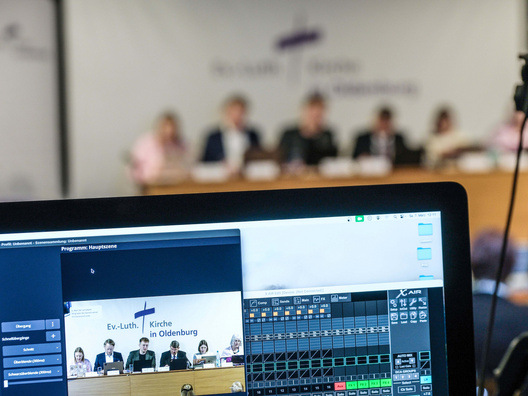 Es ist ein Bildschirm mit Menschen im Hintergrund zu sehen.