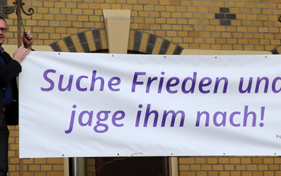 Bischof Thomas Adomeit (li.) und Birgit Carmona Scheider vom Bischofsbüro befestigen das Banner mit den Psalmworten „Suche Frieden und jage ihm nach!“ (Psalm 34,15) am Gebäude des Oberkirchenrates im Oldenburger Philosophenweg. Foto: ELKiO/D.-M. Grötzsch