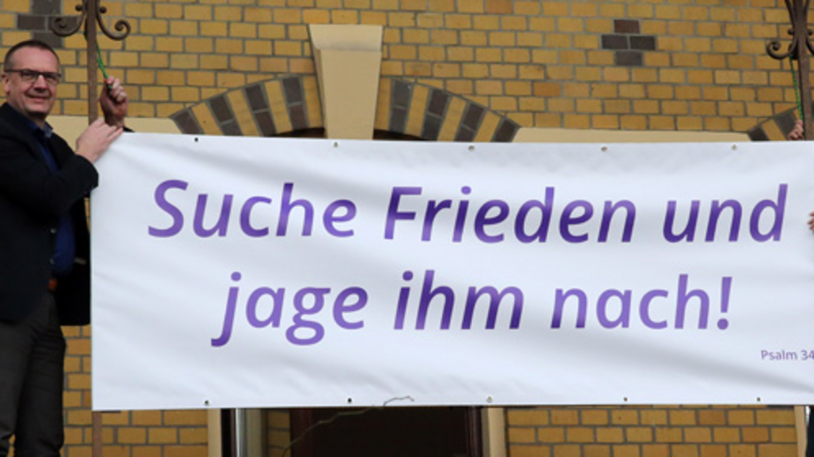 Bischof Thomas Adomeit (li.) und Birgit Carmona Scheider vom Bischofsbüro befestigen das Banner mit den Psalmworten „Suche Frieden und jage ihm nach!“ (Psalm 34,15) am Gebäude des Oberkirchenrates im Oldenburger Philosophenweg. Foto: ELKiO/D.-M. Grötzsch