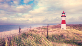 Ein Strand mit Meer im Hintergrund und einem strahlenden Leuchtturm auf der rechten Bildseite.