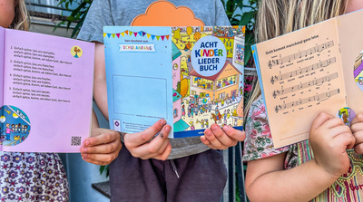 Drei Kinder halten das "AchtKinderliederbuch" in die Kamera