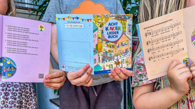 Drei Kinder halten das "AchtKinderliederbuch" in die Kamera