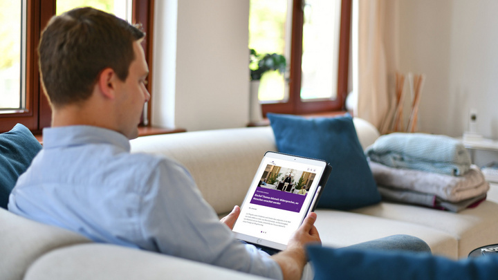 Mann sitzt auf einem Sofa und liest einen Newsletter auf einem Tablet.