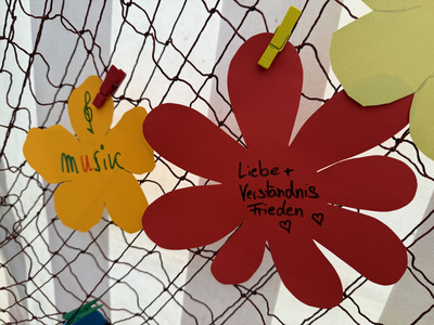 Bunte Pappblüten hängen an einem Fischernetz, darauf stehen Dinge wie "Musik" und "Frieden"