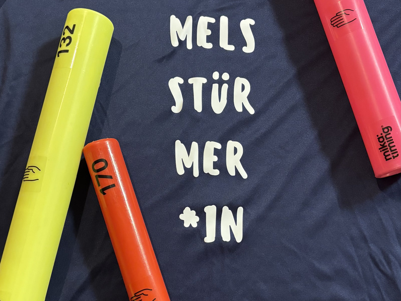 Blaues "Himmelsstürmer"-Shirt, darauf drei bunte Staffelstäbe