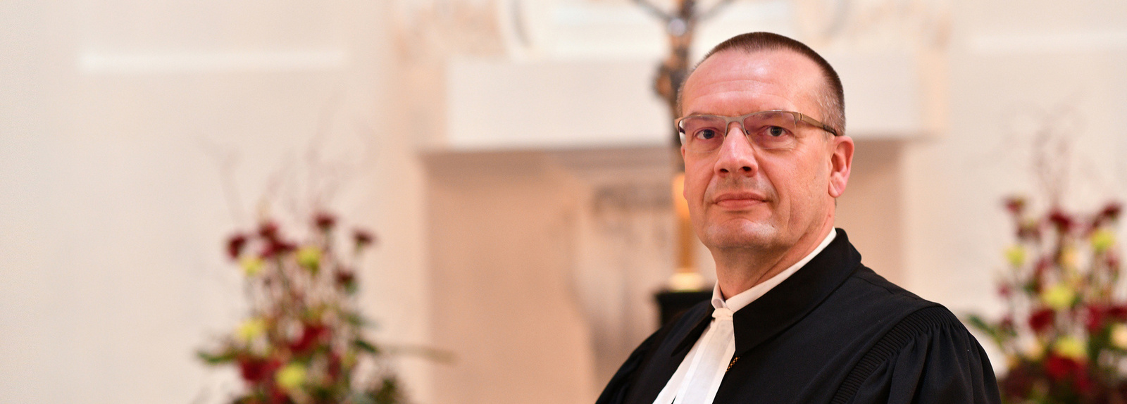 Bischof Thomas Adomeit im Talar schaut in die Kamera. Im hellen Hintergrund ist verschwommen ein Altar und ein Kreuz zu sehen.