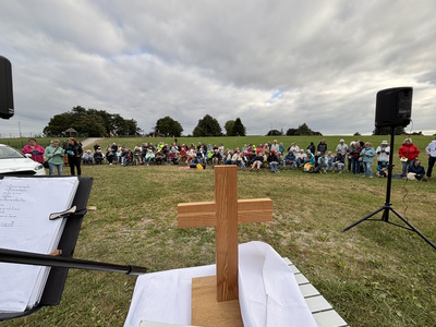 Kirche-am-Deich-2026_Strand-Campingplatz.jpg Im Vordergrund steht ein Altar-Holzkreuz, im Hintergrund sitzen viele Teilnehmende auf Stühlen im grünen Gras.