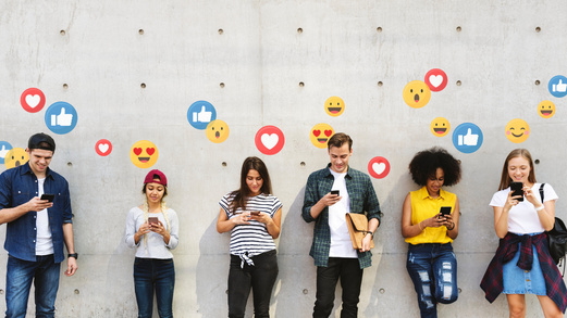 Junge Menschen stehen vor einer Wand. Sie schauen auf ihre Smartphones. An der Wand befinden sich bunte Symbole mit Herzen und Smileys.