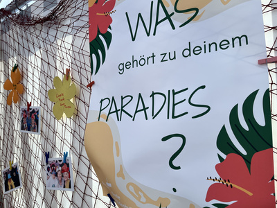Banner mit dem Schriftzug "Was gehört zu deinem Paradies?", im Hintergrund hängen Bilder aus der Fotobox aufgehängt an einem Fischernetz.